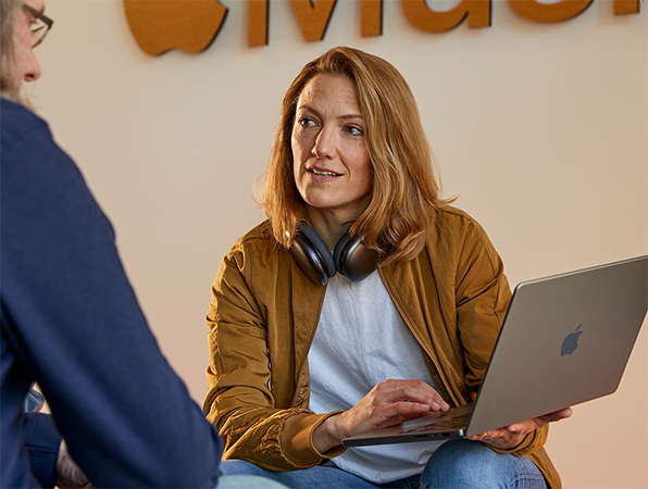 Zwei Apple Sales und Business Development Teammitglieder im Gespräch, eine Person hält ein MacBook und sitzt vor einer Wand mit dem Apple Music Logo darauf.