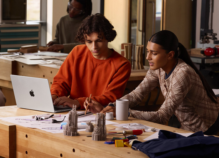 Two people collaborating on a project at a table, one person is typing on a MacBook, the other is using a pencil and a ruler on paper, surrounded by art supplies.
