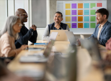 Several university staff talk around a table, MacBook open for each and a project board on the wall