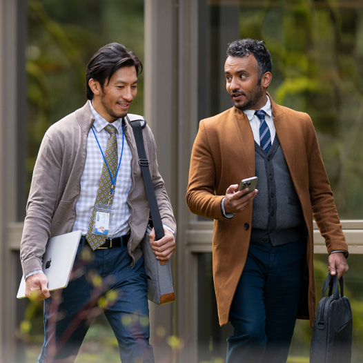 Two colleagues walk and talk outdoors, one carrying a MacBook while the other references something on their iPhone