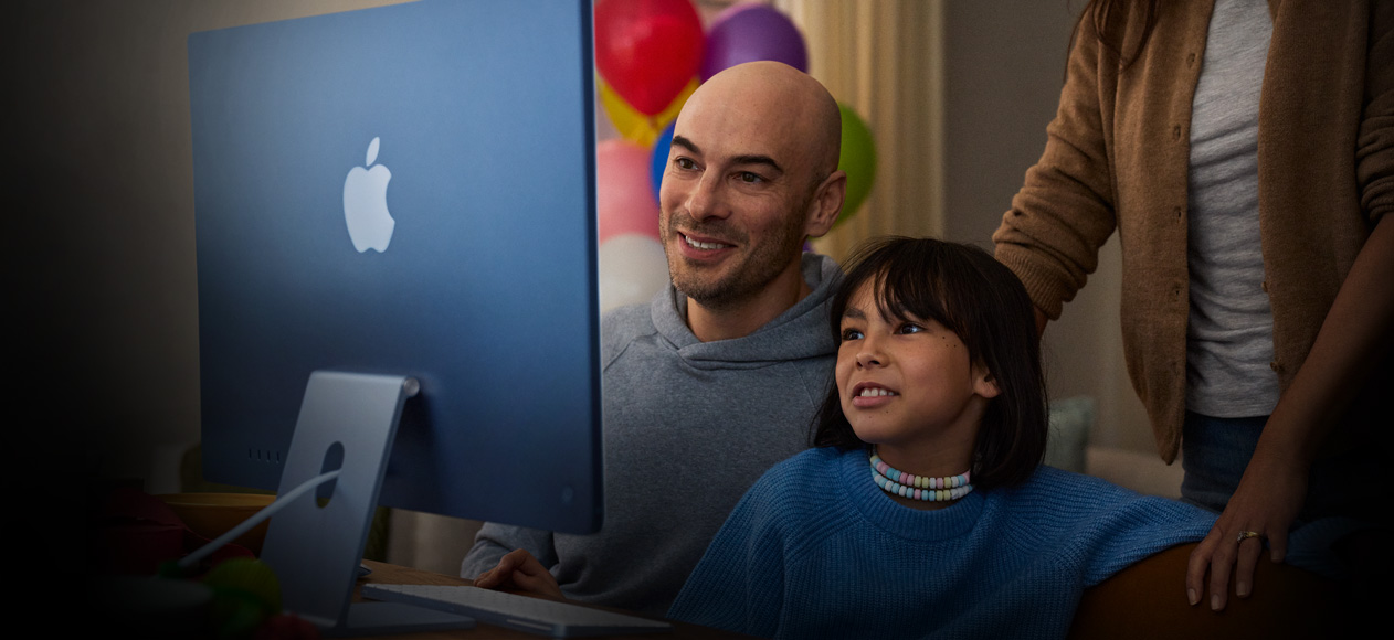 Dva lidé sedí vedle sebe a&nbsp;používají iMac, zatímco třetí člověk stojí za nimi a&nbsp;dívá se.